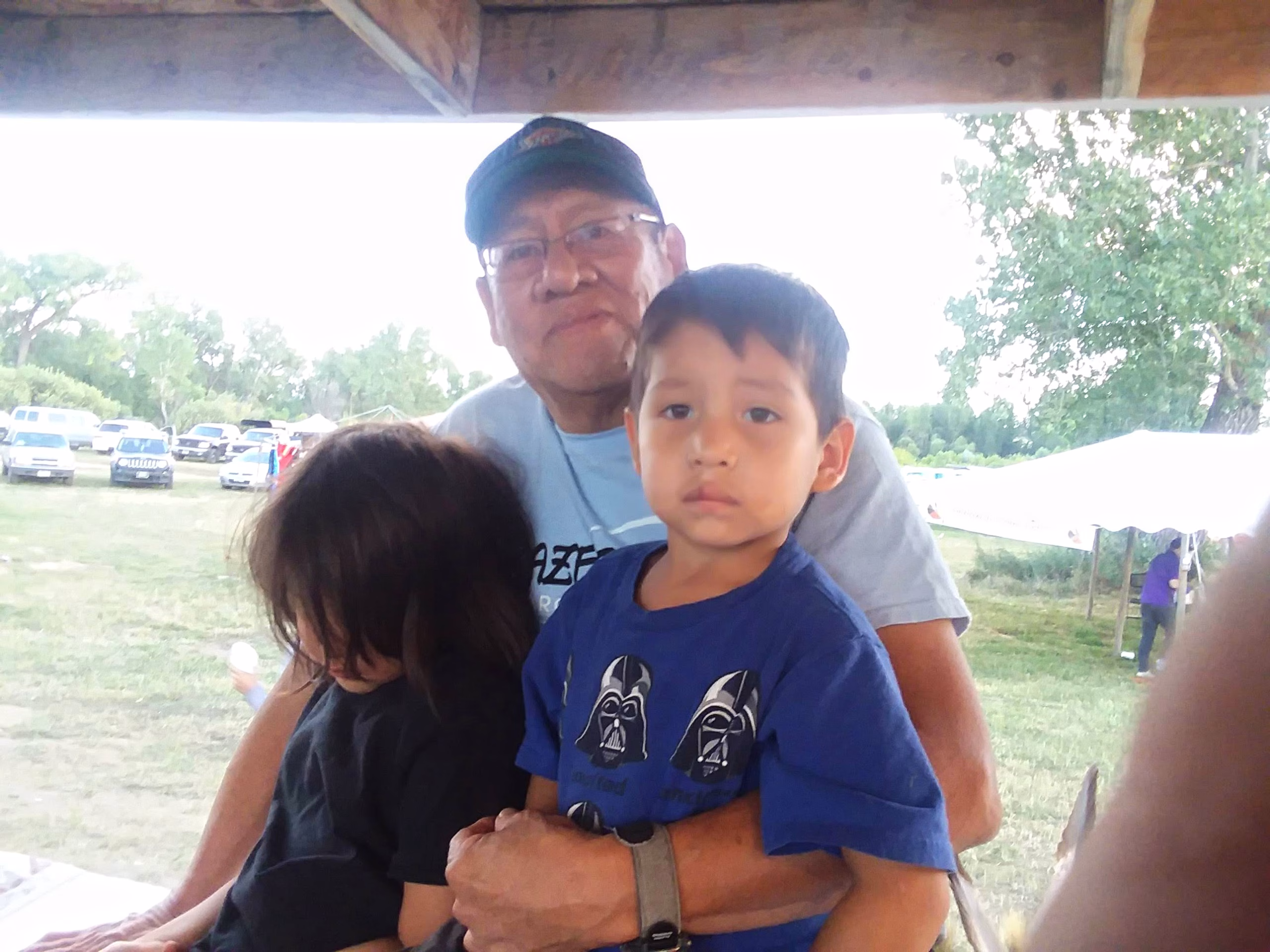 An older man wearing glasses and a cap sits outdoors, holding two young children; one child faces the camera while the other looks down, with trees and parked cars in the background.